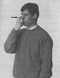 Esimerkki 5. Mustavalkoinen kuva jossa tummahiuksinen mies tummassa villapaidassa jonka alta näkyy valkoiset kaulukset pitää oikean käden etu- ja keskisormella nenästään kiinni pienen mustan nuolen osoittaessa sormia.