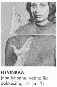 Esimerkki 27. Mustavalkoinen kuva tummahiuksisesta naisesta tummassa neuleessa näyttämässä OK-merkkiä (saman käden peukalon ja etusormen päät koskettavat toisiaan) ja alemmassa kuvassa kämmen auki ylöspäin suunnattuna lähellä rintaa.
