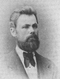 Kuva 5. Lorenz Eklund (1847–1892). – Kuurojen Liitto ry, Kuurojen museo. Mustavalkoinen rintakuva parrakkaasta tummahiuksisesta miehestä jolla valkoinen kauluspaita, tumma puvuntakki ja rusetti.