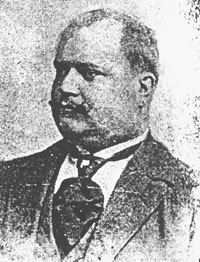 Kuva 6. Julius Hirn (1868–1914). – Kuurojen Liitto ry, Kuurojen museo. Mustavalkoinen rintakuva pyöreähköstä vanhemmasta miehestä jolla viikset ja tumma puku leveällä kravaatilla.